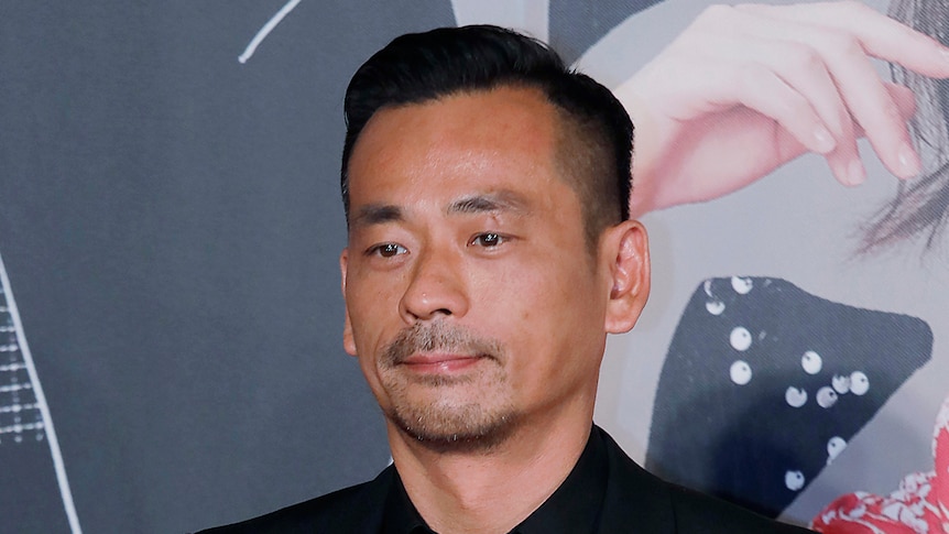 A man in a black shirt, suit and tie stands at a movie premiere 