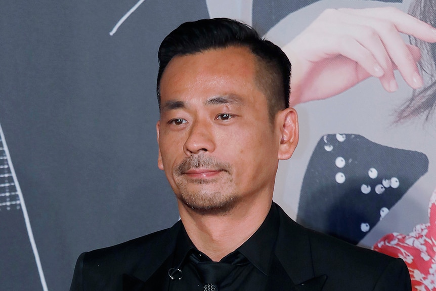 A Chinese man in a black shirt, suit and tie stands at a movie premiere 