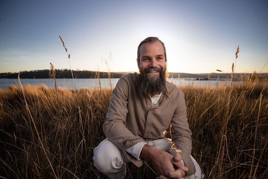 A bearded man wears khaki pants and button up while crouching in dry grass. A large river can be seen in background