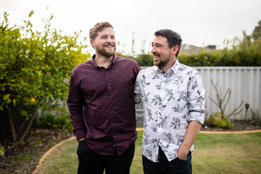 Two caucasian men in button-down shirts smile in a suburban backyard.