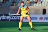Woman dressed in hockey uniform stands poised with hockey sit in the air, ready to hit ball.