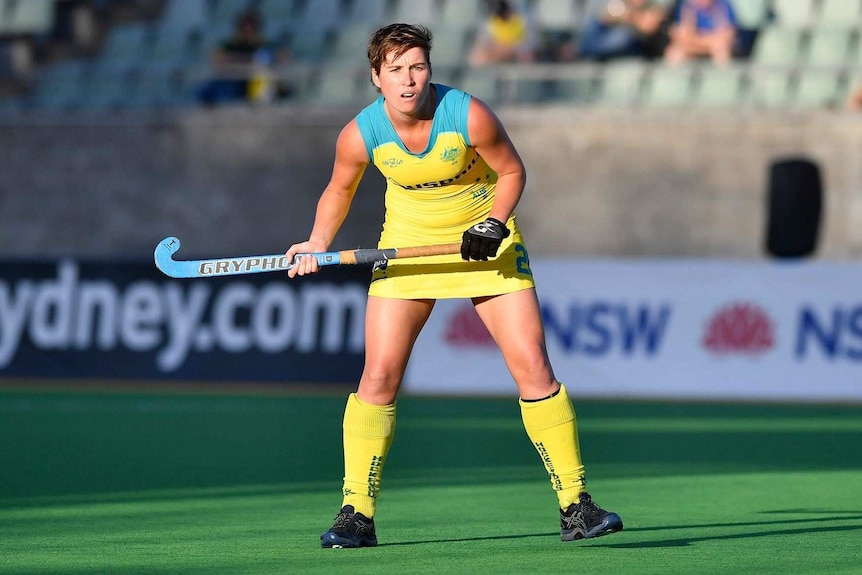 Woman dressed in hockey uniform stands poised with hockey sit in the air, ready to hit ball.