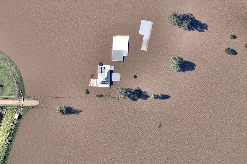 Aerial view of a dirt road and three buildings submerged in brown flood water.