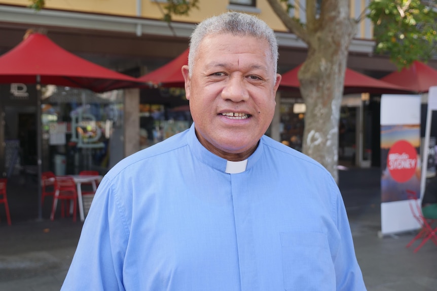 A man wearing a blue shirt with a clerical collar smiles