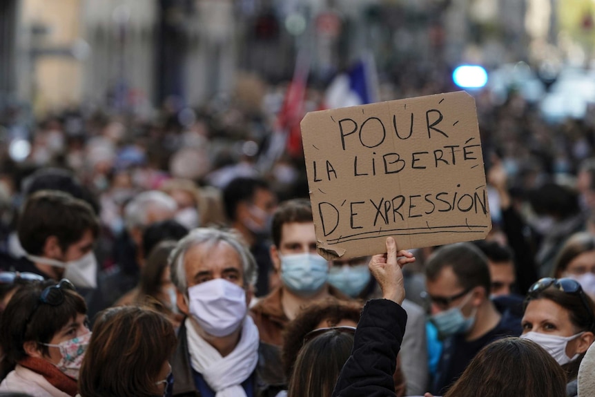 A cardboard sign carrying the words 'for the freedom of expression' in French is held above a crowd of masked demonstrators.