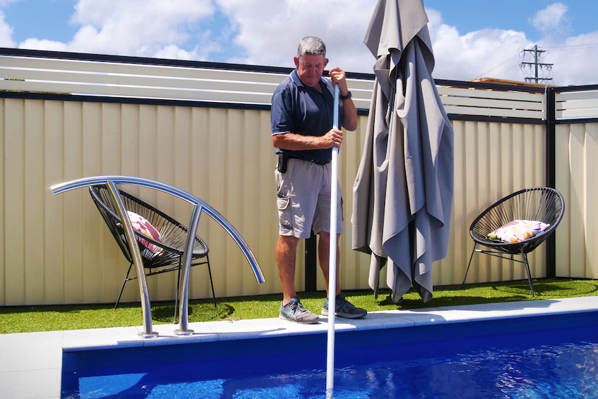 A man in a work uniform cleaning a backyard swimming pool. 