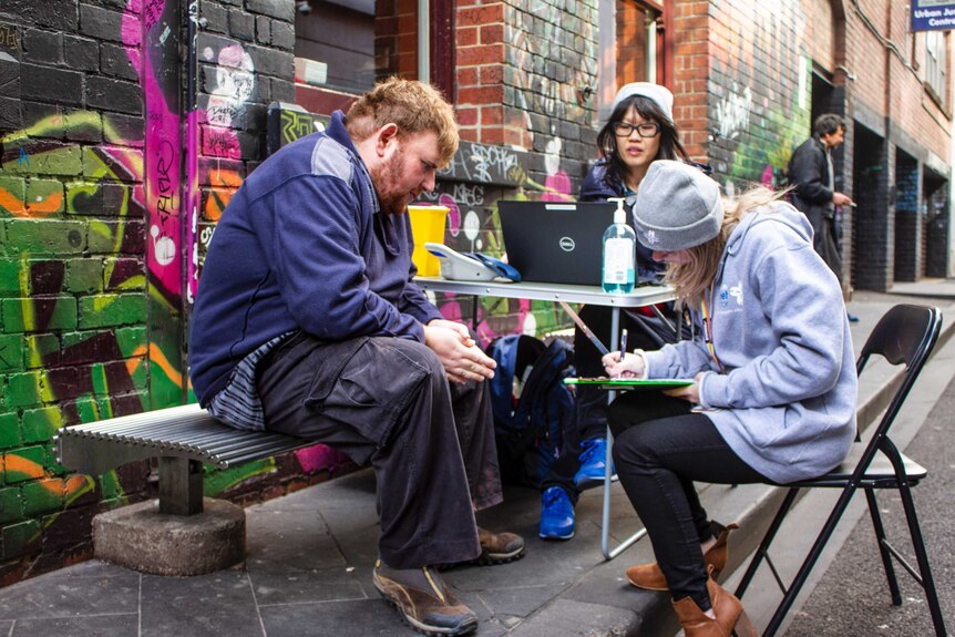 A man and two women sit around a fold up table in an alley way, one woman takes notes.