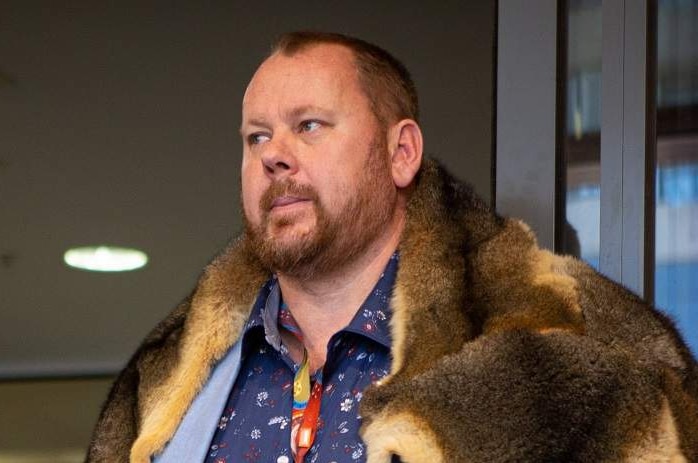 Troy Williamson stands in an indoors corridor, wearing a possum-skin cloak over his shirt and jacket.