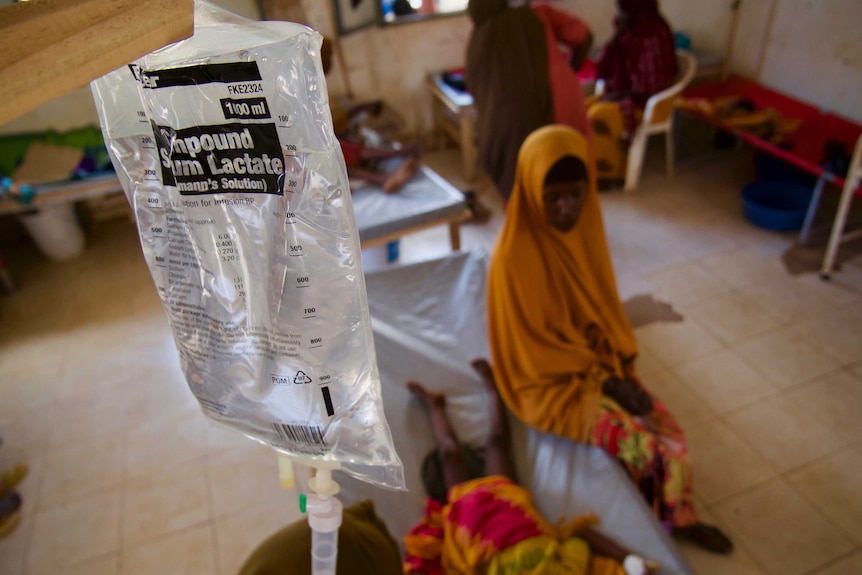 Woman sits on the side of a child's hospital bed. An IV fluid drip hanging in foreground.