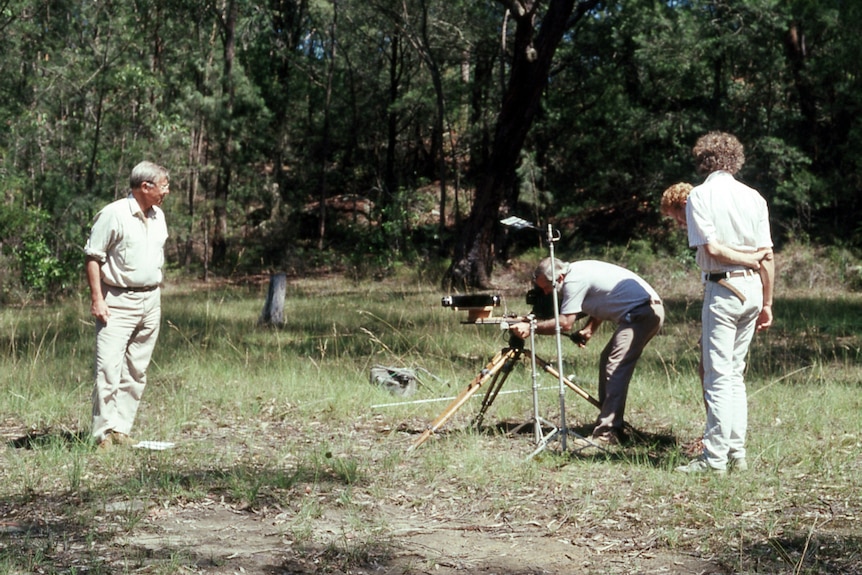 A man in a bush area bends down behind a video camera, with another man standing in front of the camera.