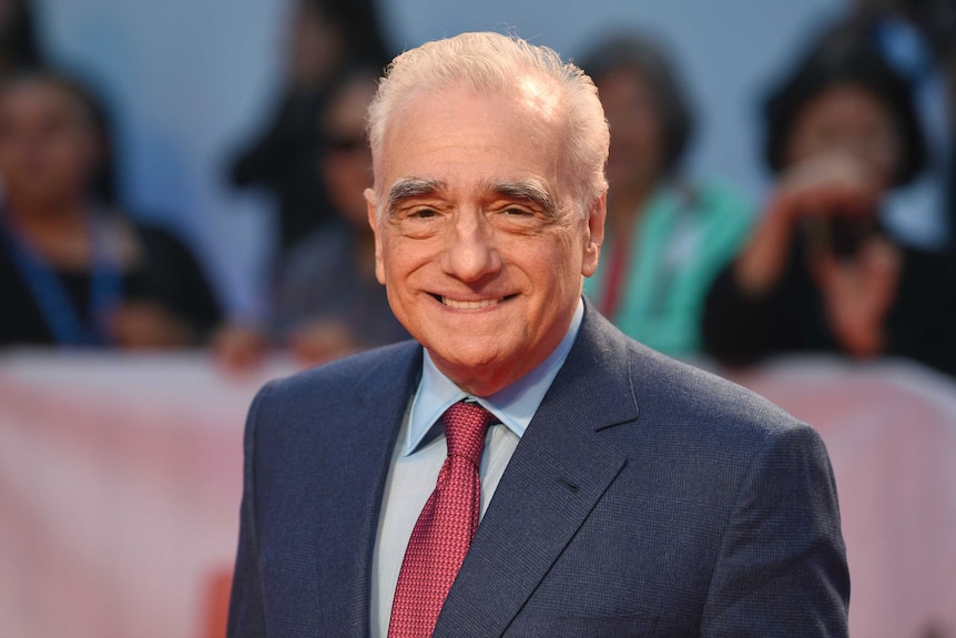 Martin Scorsese smiles in the direction of the camera while standing on a red carpet.