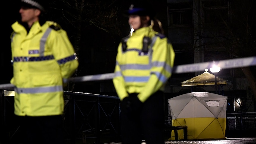 Police officers stand guard in front of a cordoned-off area with a police tent.