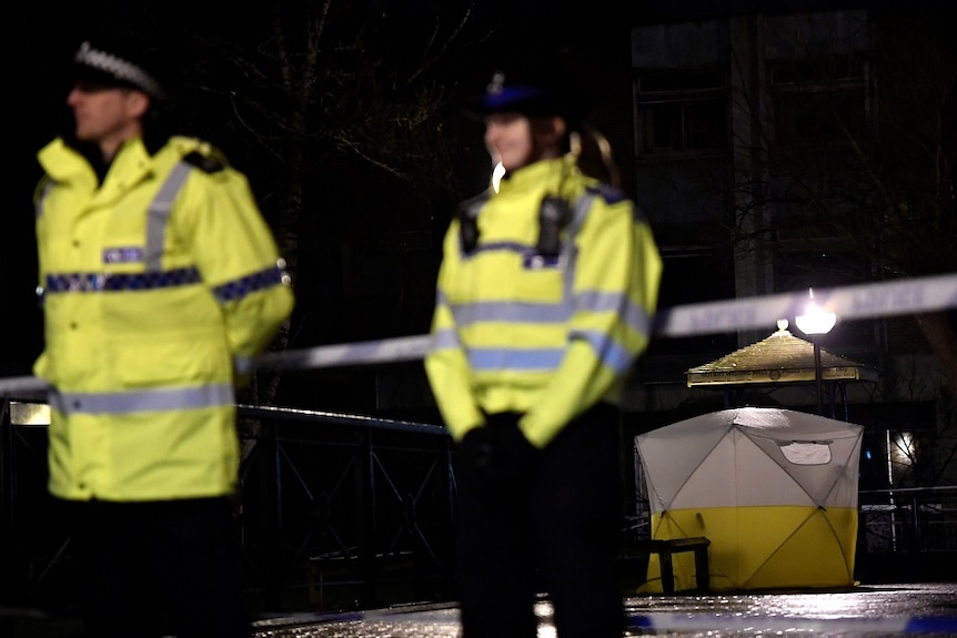 Police officers stand guard in front of a cordoned-off area with a police tent.