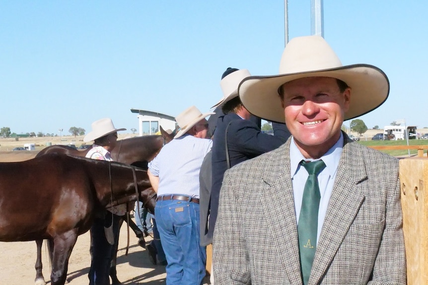 Matthew Holz stands in a tweed jacket and white hat, leaning against a wooden rail with horses and people standing behind him.