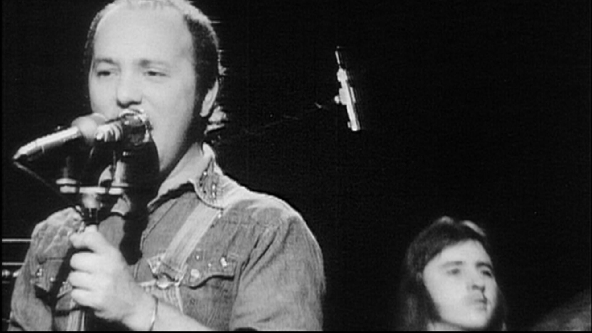 A black and white photo of a man in a denim jacke singing into a microphone with a long haired man seated behind him.