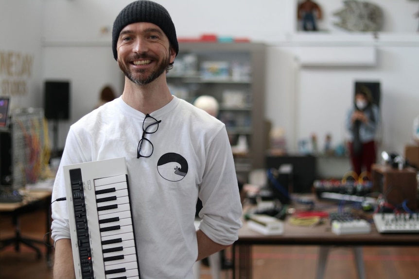 A smiling man in a white shirt and a black beanie holds a keyboard.