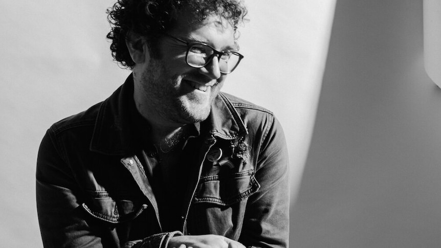 black and white portrait of smiling man, looking down, hands clasped in front of him. He has curly hair and spectacles.