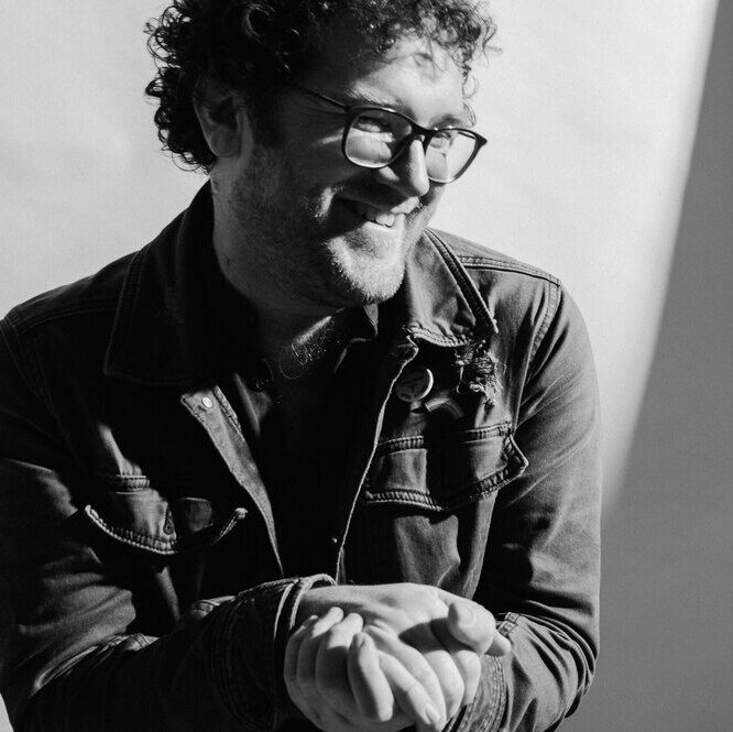 black and white portrait of smiling man, looking down, hands clasped in front of him. He has curly hair and spectacles.