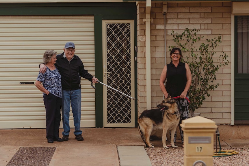 An elderly man and woman and a middle aged woman with a dog stand in front of a house