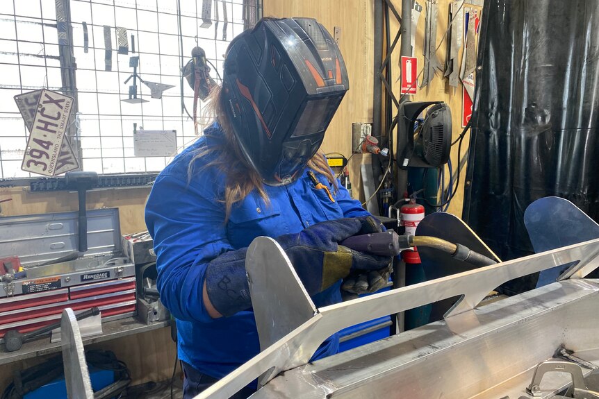 A person with long blond hair wears a protective helmet, gloves and blue shirt welds.