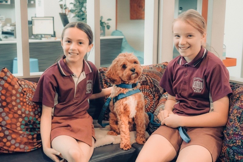 Young students Bonnie and Milla smile, wearing their uniforms and smiling on the couch with Monty the cavoodle.