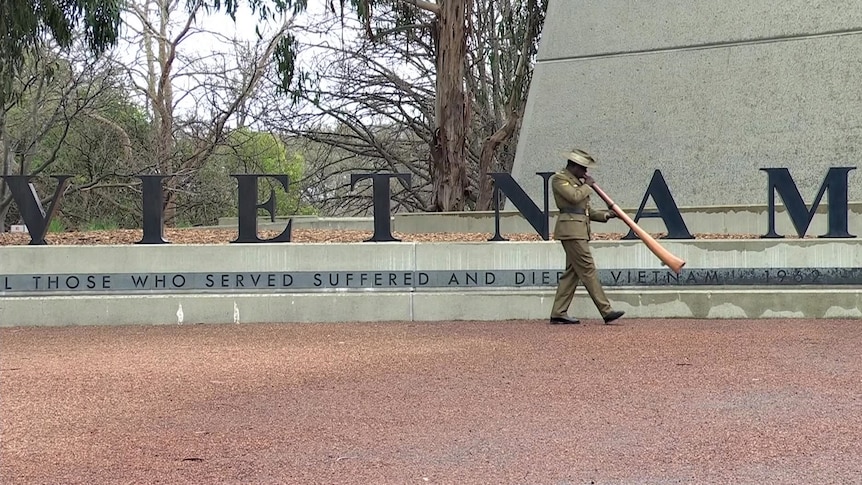 A man plays didgeridoo in front of the memorial, which says VIETNAM.