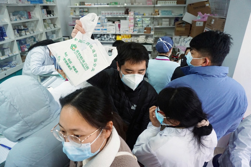 A man emerges from a crowd of people lining up at a chemist counter, holding a plastic bag aloft