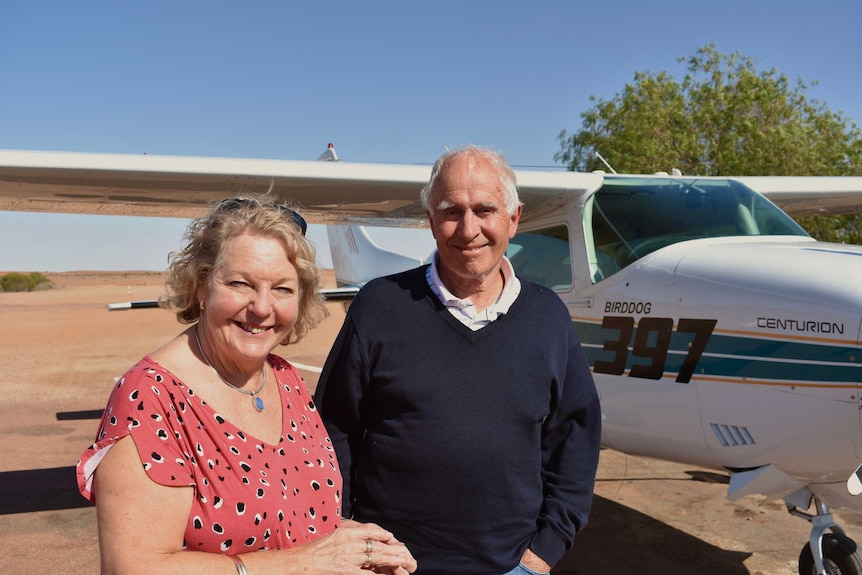 An older couple stand in front of a plane.  Both are smiling and looking at the camera.