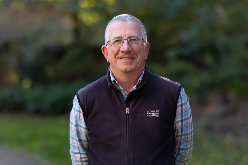 Middle aged man in plaid shirt and fleece vest, wearing glassed smiles at camera 