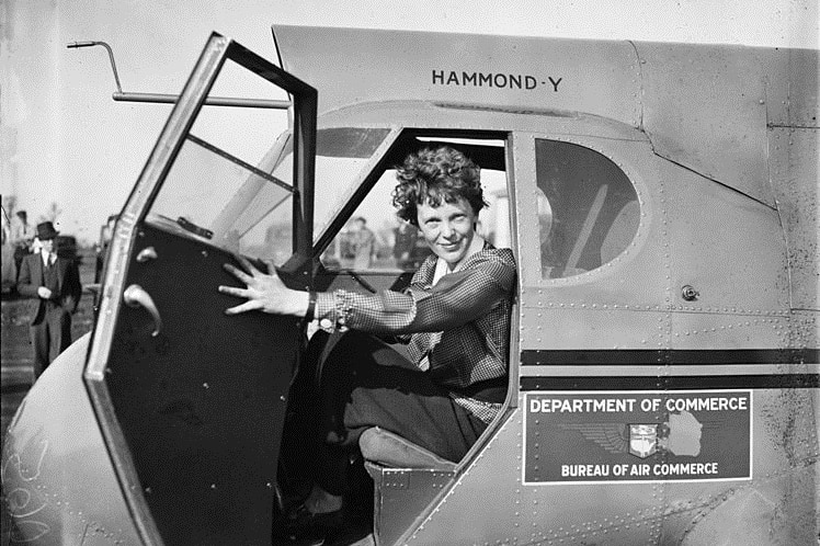 Amelia Earhart sits in the cockpit of a plane, with the door open, she is looking at the camera in this 1936 photograph.