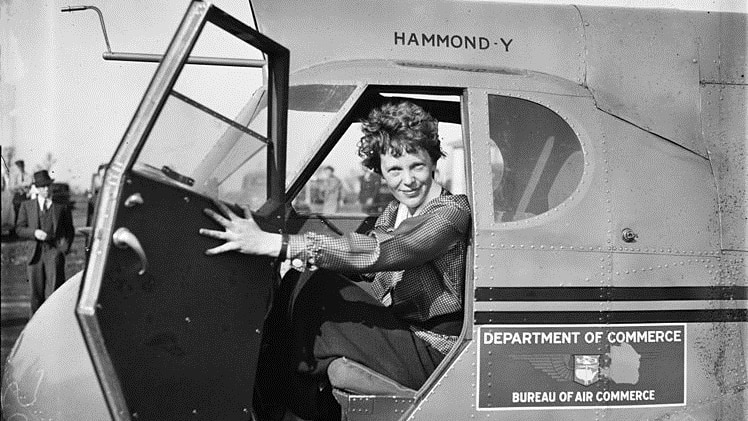 Amelia Earhart sits in the cockpit of a plane, with the door open, she is looking at the camera in this 1936 photograph.