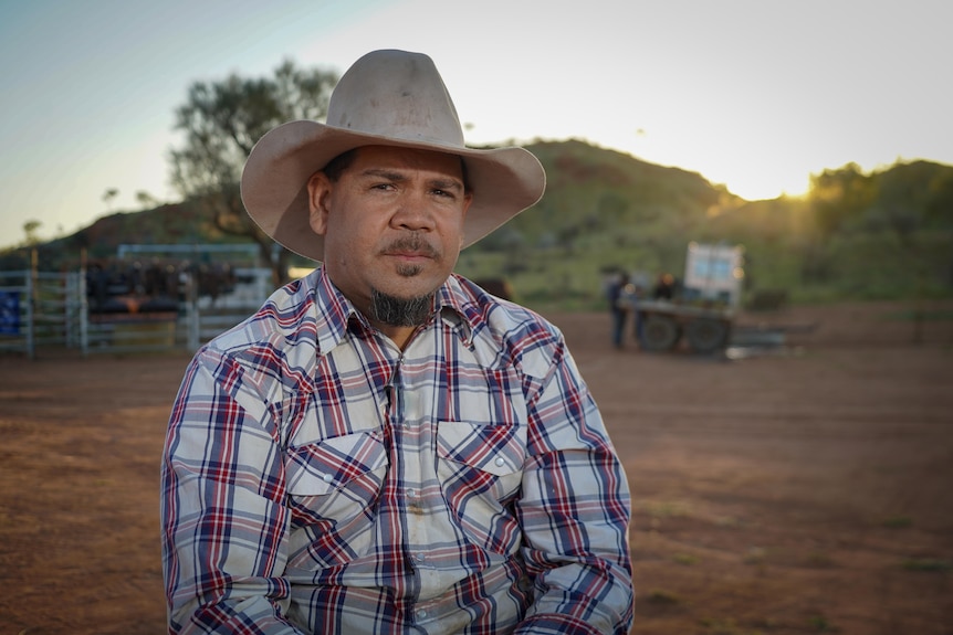 A man wearing a cowboy hat and a checked shirt looks seriously at the camera.