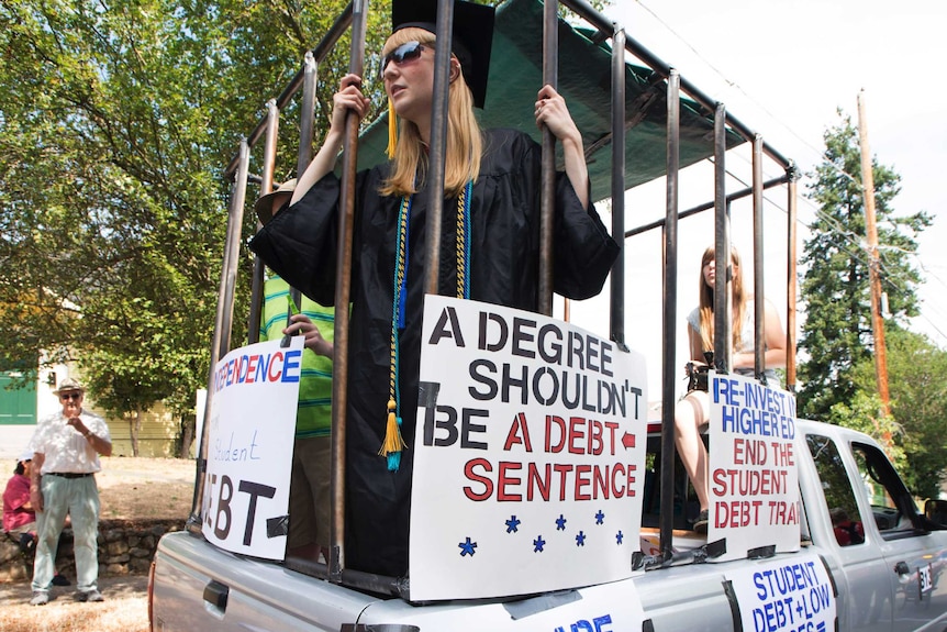 A girl in a graduate gown holds the bars of a cage with protest signs about student debt