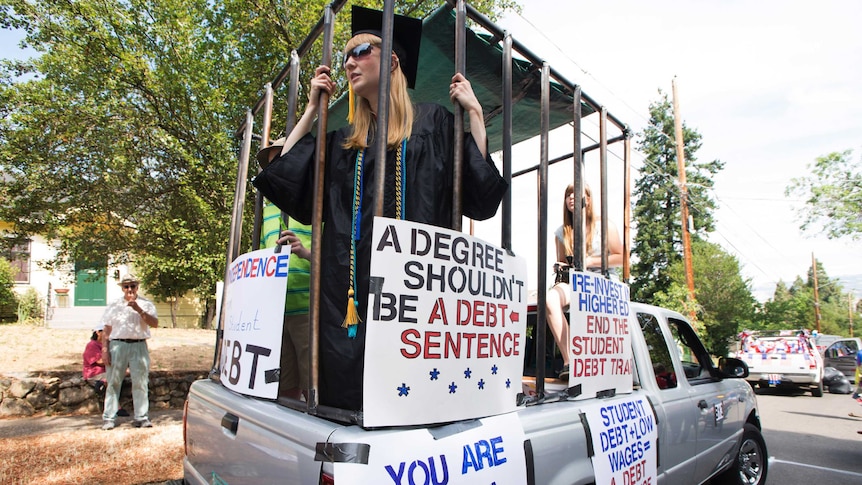 A girl in a graduate gown holds the bars of a cage with protest signs about student debt