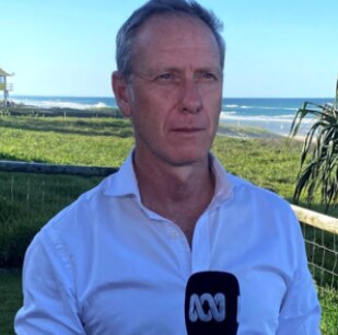 A man (David Mark) standing outside with the beach in the background holding a microphone with the ABC logo.