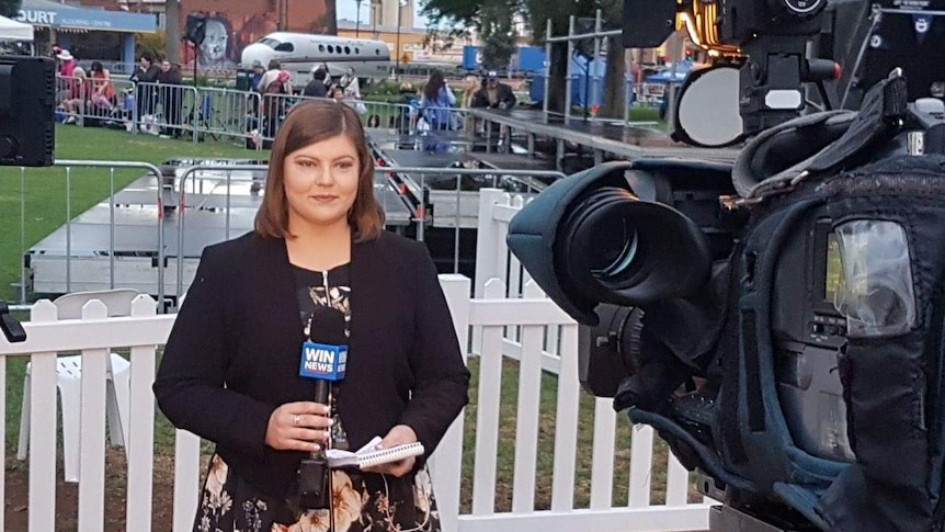 A female reporter hold a WIN News microphone and a notepad reports in front of a camera.