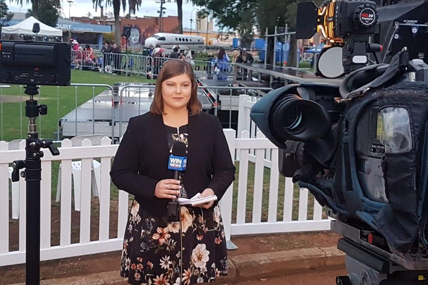 A female reporter hold a WIN News microphone and a notepad reports in front of a camera.