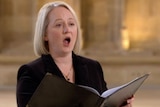 A blond woman holds a hymn book while singing in a church