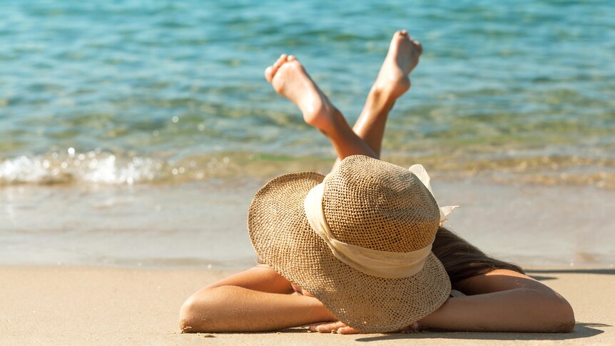 A person wearing a sunhat lies on the beach near the water.