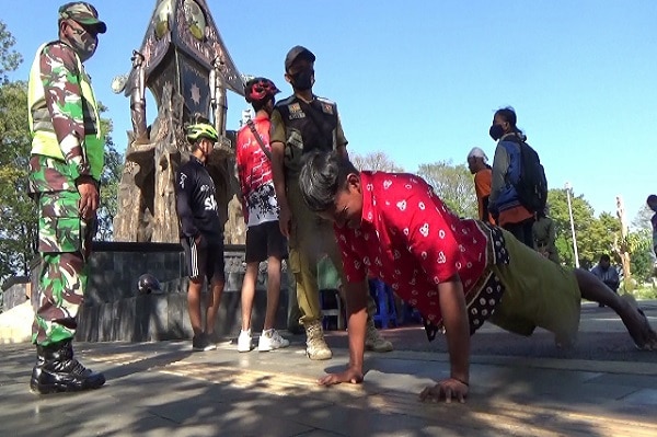 A man doing push-ups on a street in Indonesia is being watched by two officials wearing masks.