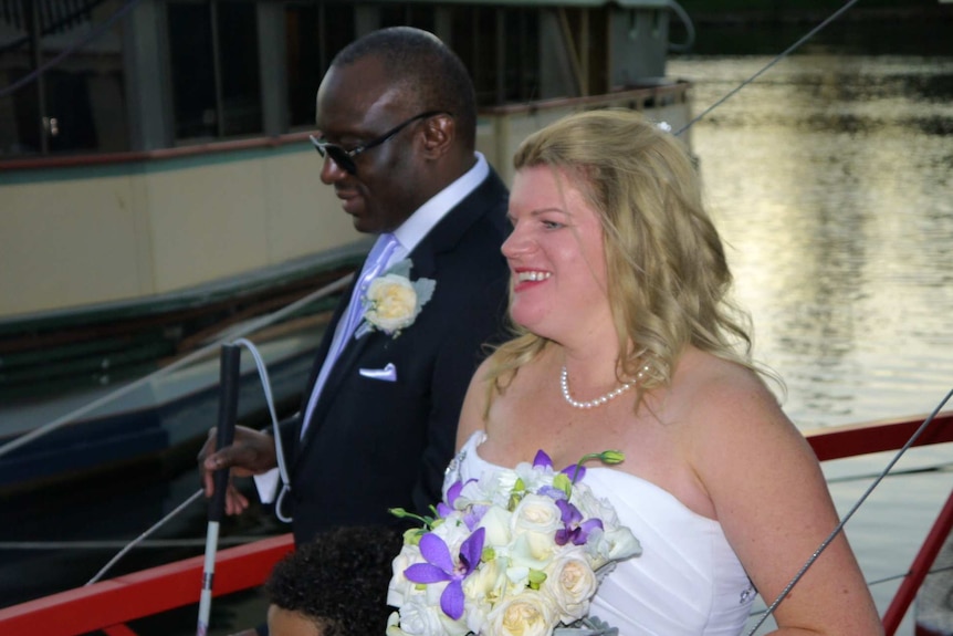 Krystal, wearing a wedding dress, and Nemoy, wearing a tuxedo, walk together next to a lake. Krystal carries a bouquet.