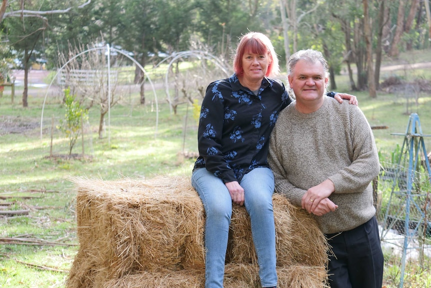 A women is sitting on top of a hay bale learning on her husband standing next to her looking at the camera.