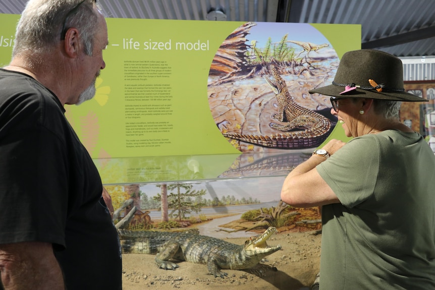 A man in a black shirt and a woman in a green shirt with a brown cowboy hat stand in front of a museum cabinet.