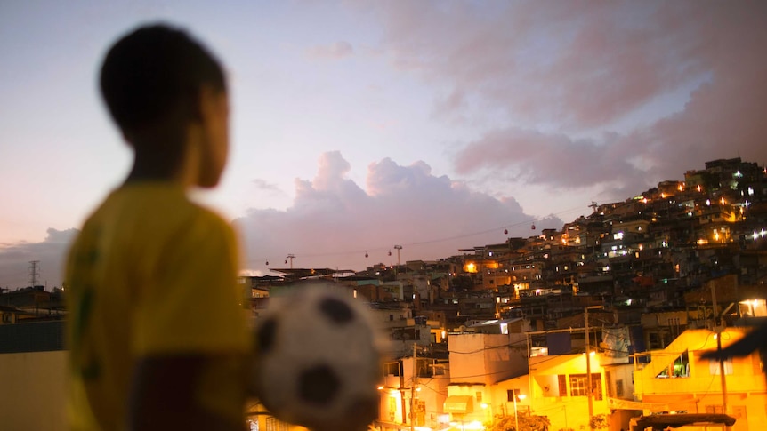 A young Brazilian footballer in yellow football shirt with a ball, overlooking a favela in Brazil.