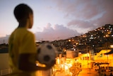 A young Brazilian footballer in yellow football shirt with a ball, overlooking a favela in Brazil.