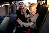 A boy sitting in a car seat doing a thumbs-up sign with a woman behind