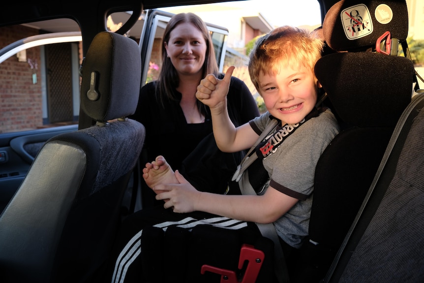 A boy sitting in a car seat doing a thumbs-up sign with a woman behind