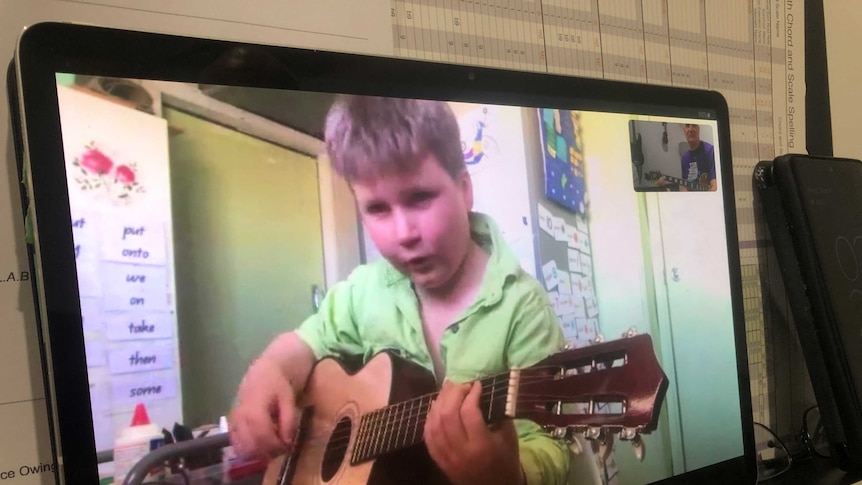 A young boy plucks tentatively at his guitar strings while his tutor watches him on screen during a remote lesson.