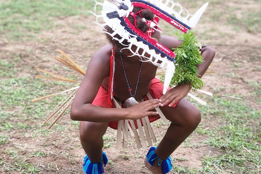 A young man wearing traditional headgear and bamboo skirt squats down with arms out in movement.