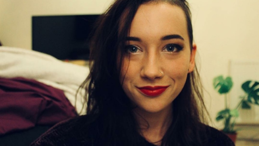 A young woman with long dark hair and red lipstick sits in her bedroom, smiling at the camera.